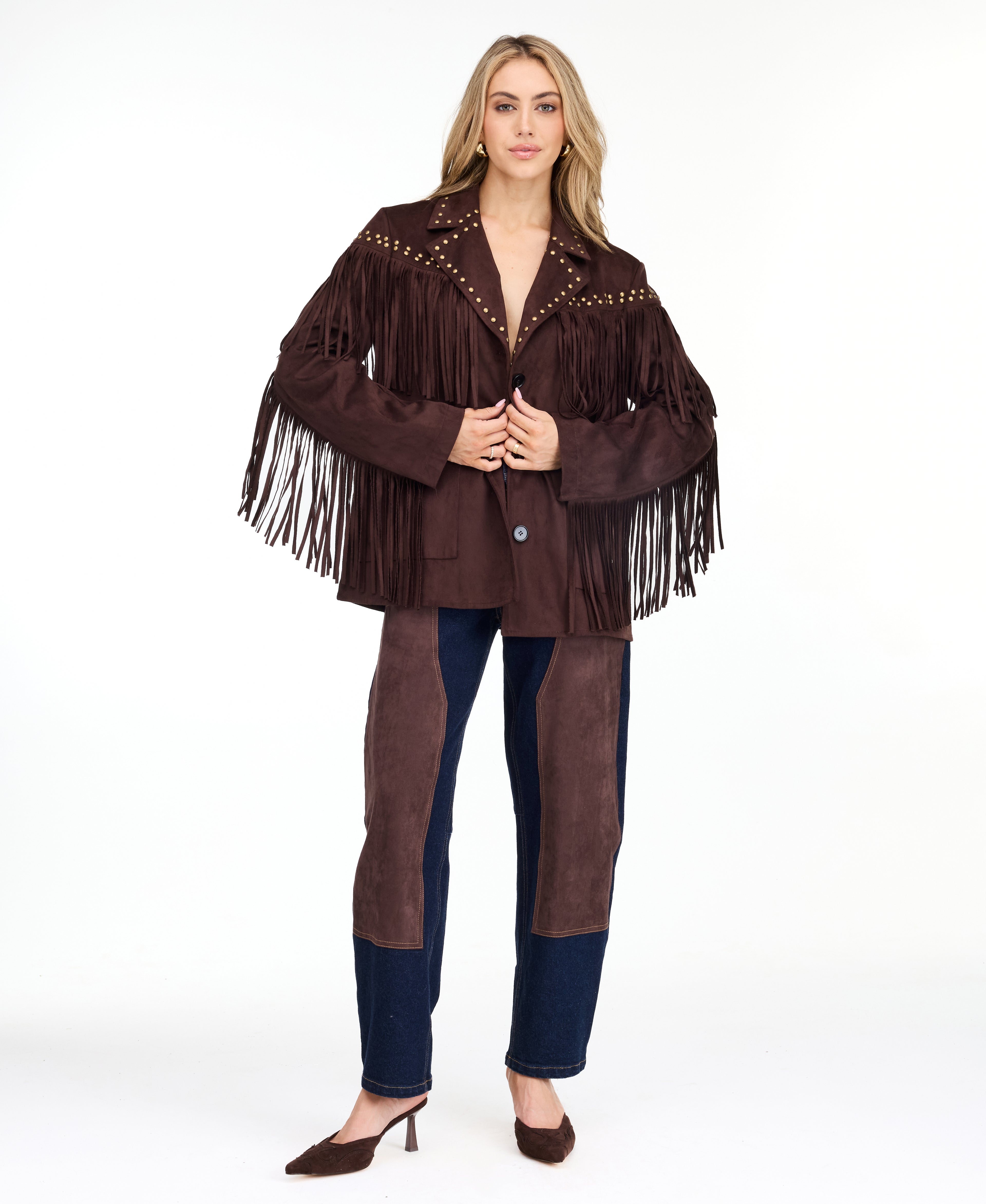 A woman modeling a dark brown suede fringe jacket with gold-tone studs and buttons, paired with brown and denim patchwork pants and brown pointed-toe heels, against a plain white studio background.