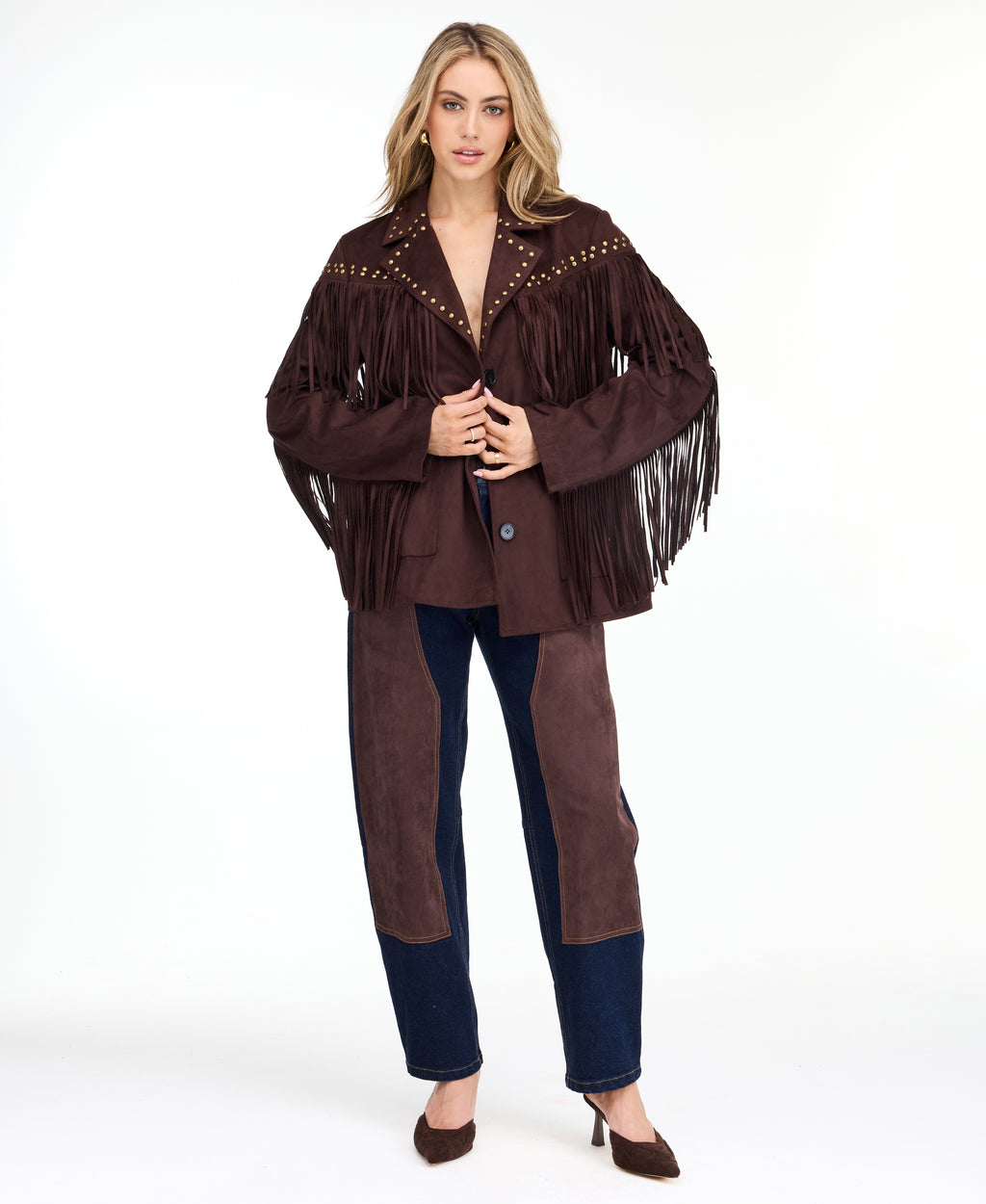 A woman modeling a dark brown fringed jacket with studded detailing, paired with dark brown and navy patchwork pants and brown heels, against a plain white studio background.