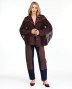 A woman modeling a dark brown fringed jacket with studded detailing, paired with dark brown and navy patchwork pants and brown heels, against a plain white studio background.