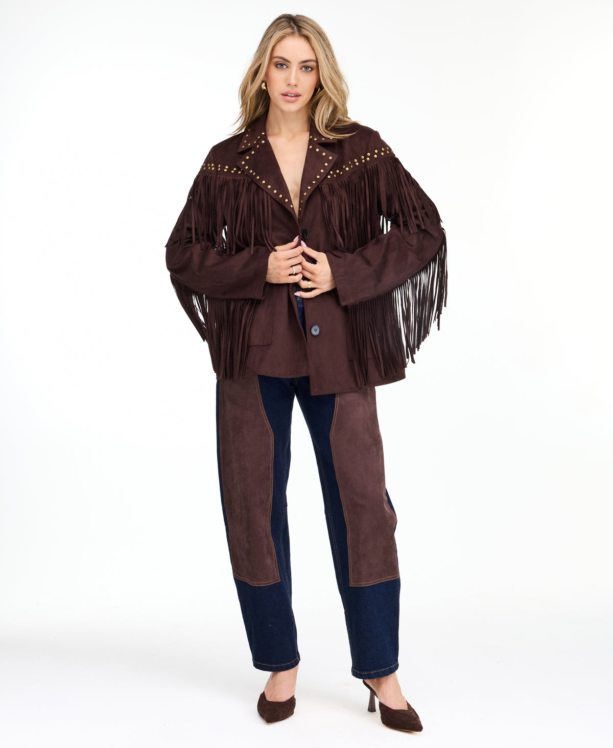 A woman modeling a dark brown fringed jacket with studded detailing, paired with dark brown and navy patchwork pants and brown heels, against a plain white studio background.