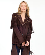 A woman modeling a dark brown suede fringe jacket with gold-tone studs and buttons, paired with brown and denim patchwork pants and brown pointed-toe heels, against a plain white studio background.