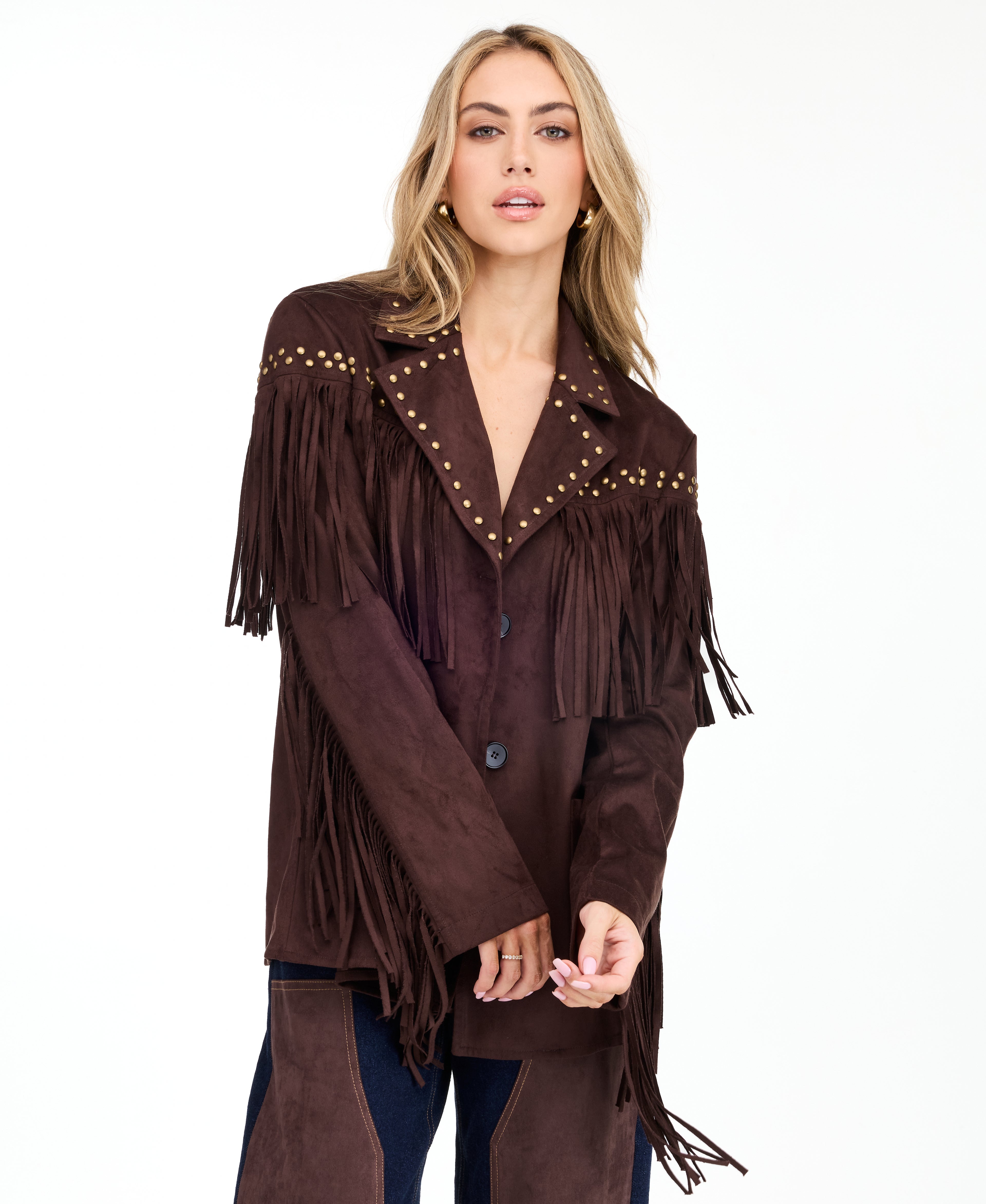 A woman modeling a dark brown suede fringe jacket with gold-tone studs and buttons, paired with brown and denim patchwork pants and brown pointed-toe heels, against a plain white studio background.