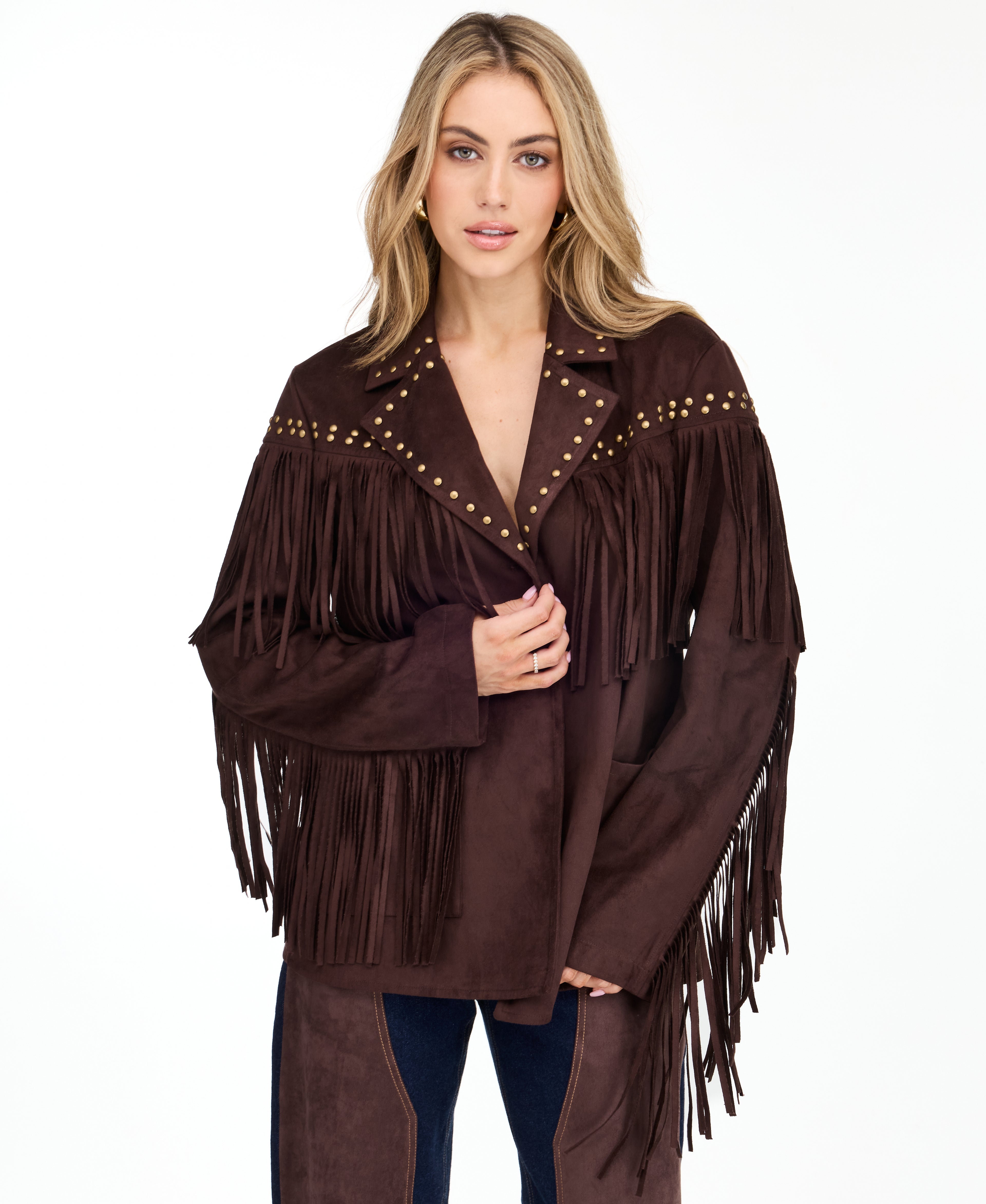 A woman modeling a dark brown suede fringe jacket with gold-tone studs and buttons, paired with brown and denim patchwork pants and brown pointed-toe heels, against a plain white studio background.