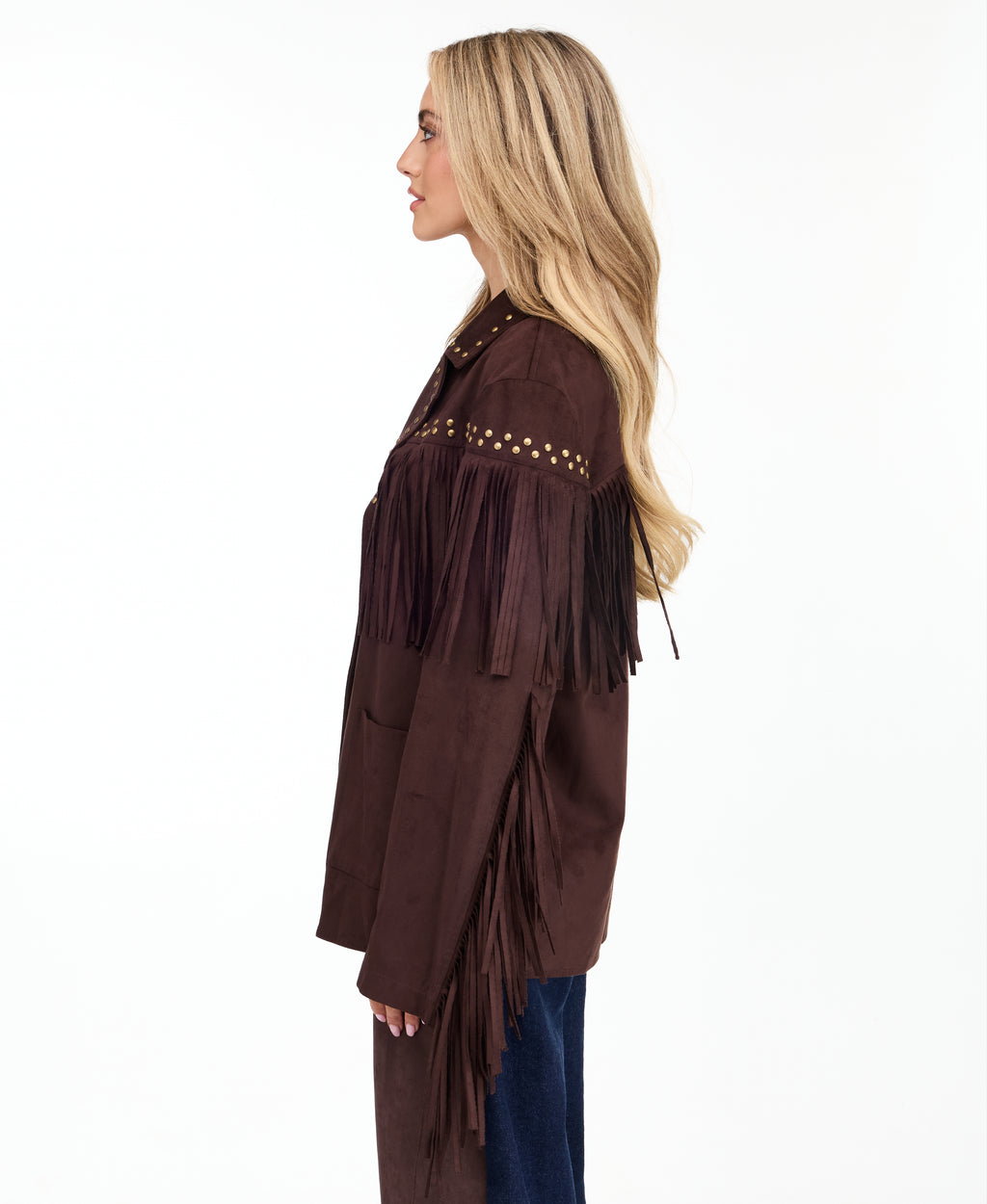 A woman modeling a dark brown suede fringe jacket with gold-tone studs and buttons, paired with brown and denim patchwork pants and brown pointed-toe heels, against a plain white studio background.