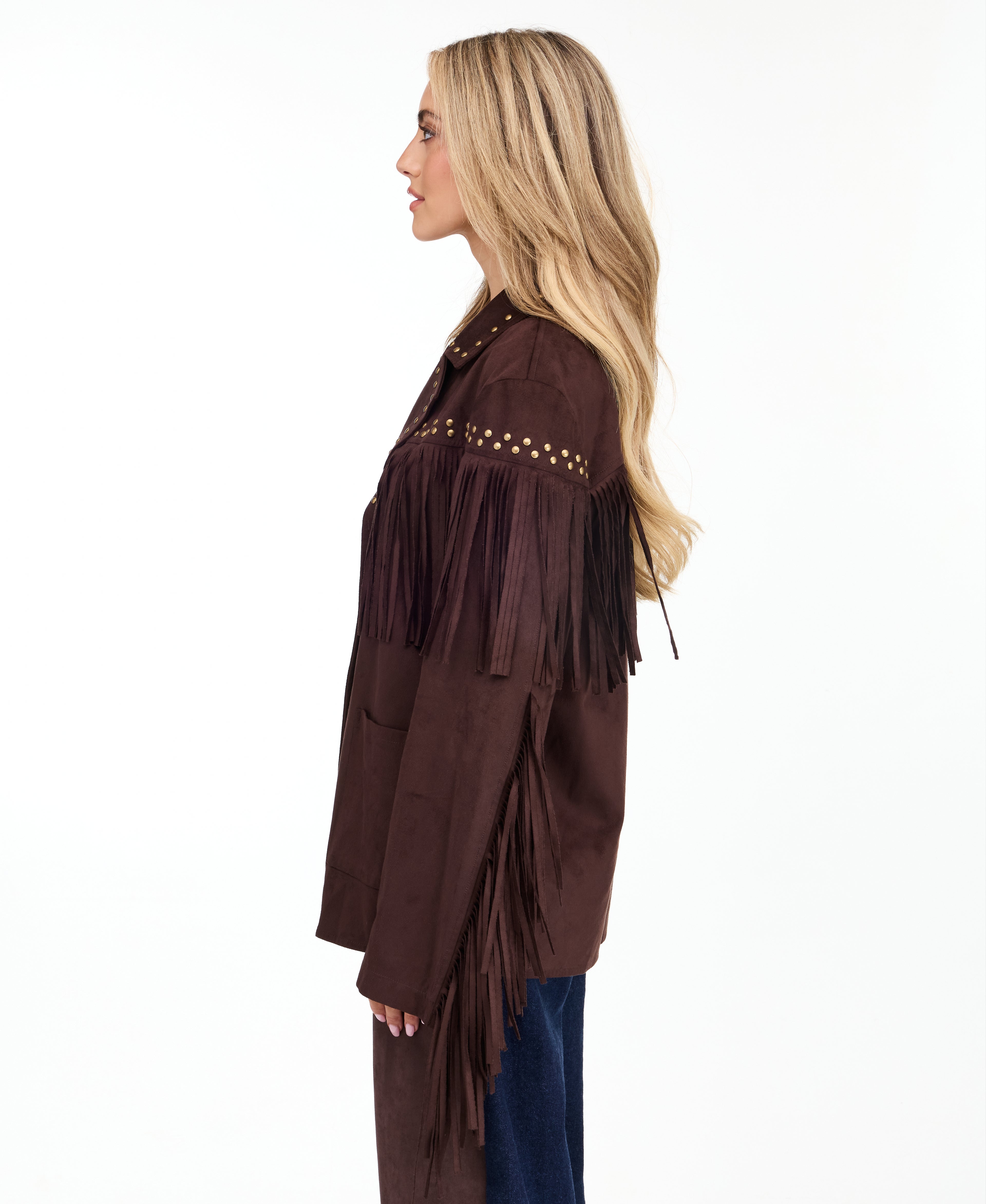 A woman modeling a dark brown suede fringe jacket with gold-tone studs and buttons, paired with brown and denim patchwork pants and brown pointed-toe heels, against a plain white studio background.