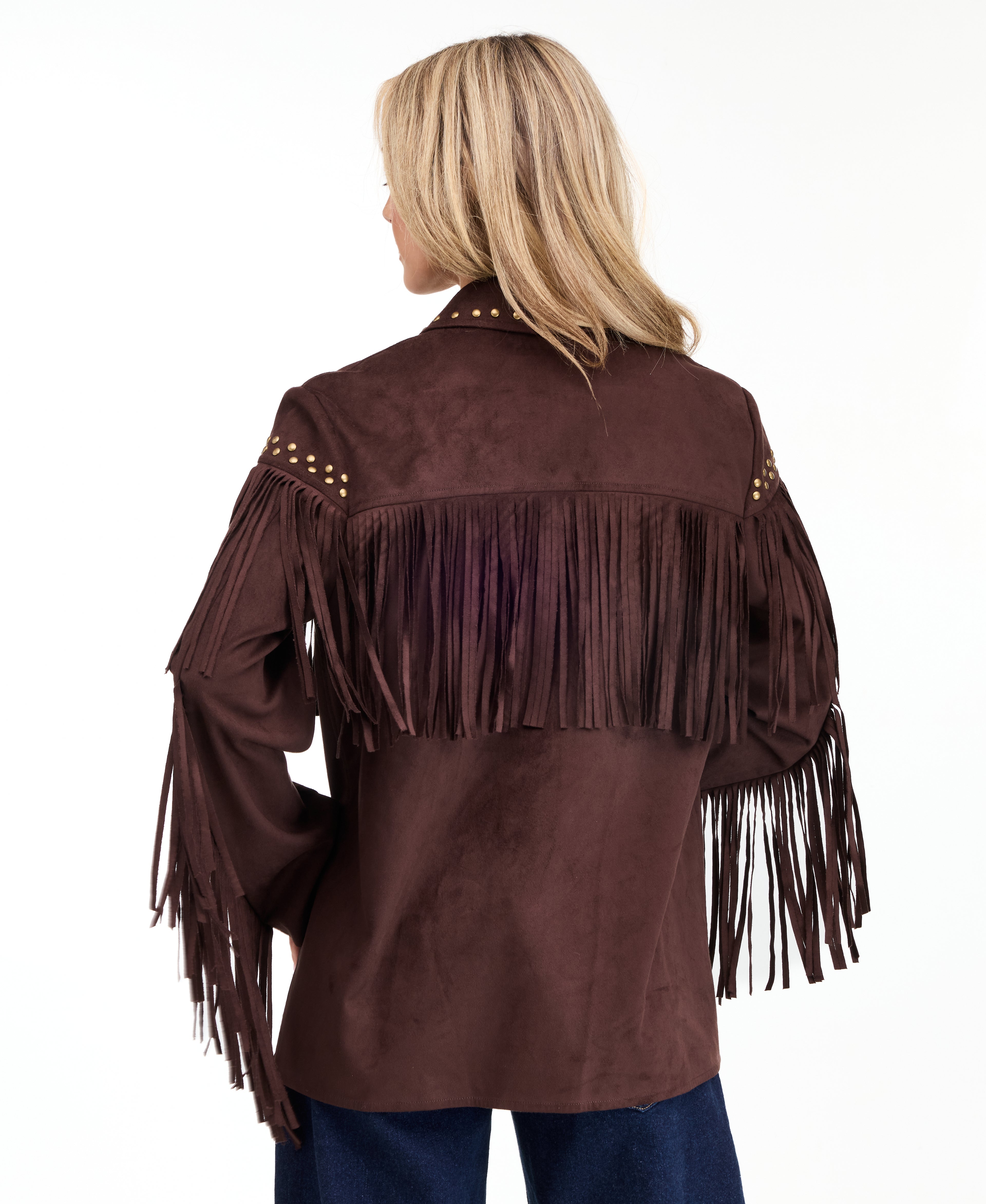 A woman modeling a dark brown suede fringe jacket with gold-tone studs and buttons, paired with brown and denim patchwork pants and brown pointed-toe heels, against a plain white studio background.