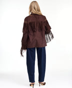 A woman modeling a dark brown fringed jacket with studded detailing, paired with dark brown and navy patchwork pants and brown heels, against a plain white studio background.