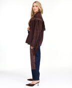 A woman modeling a dark brown suede fringe jacket with gold-tone studs and buttons, paired with brown and denim patchwork pants and brown pointed-toe heels, against a plain white studio background.