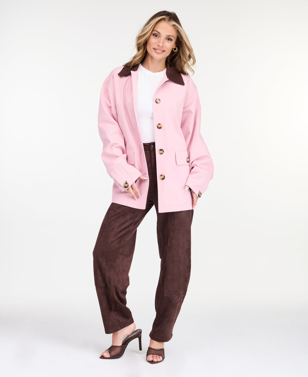A woman modeling a light pink oversized jacket with dark brown collar and buttons, worn over a white top and paired with a brown pants, on a white studio background.