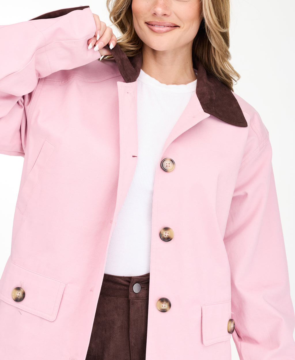 A woman modeling a light pink oversized jacket with dark brown collar and buttons, worn over a white top and paired with a brown pants, on a white studio background.