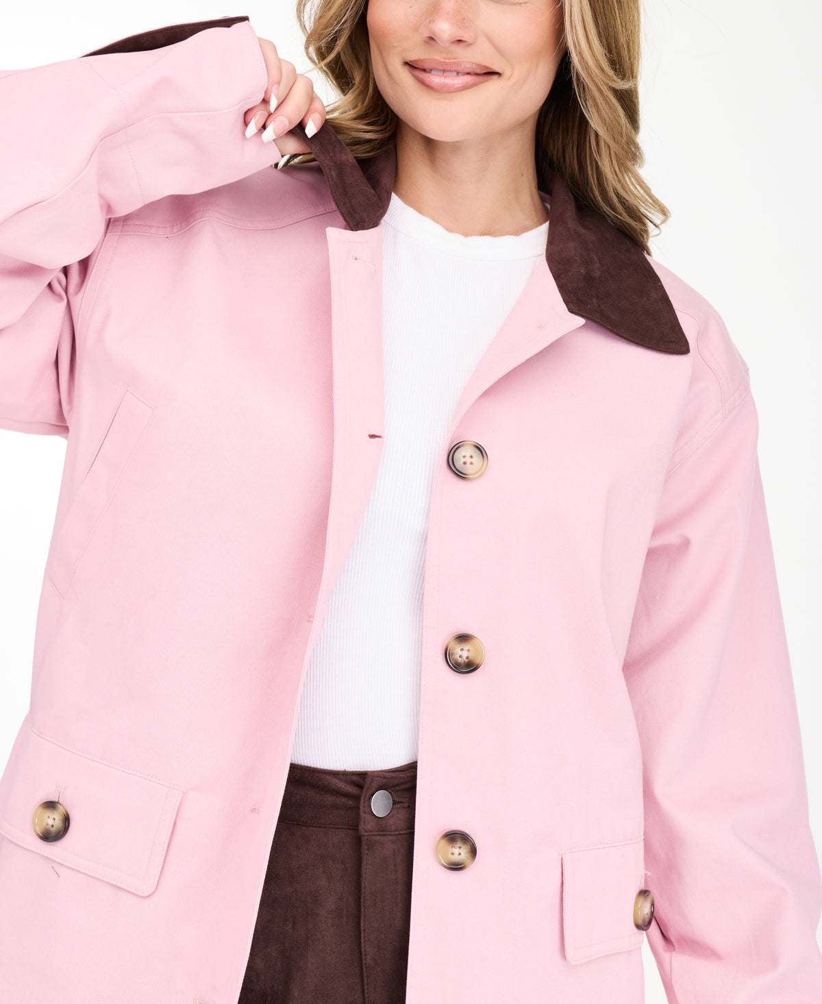 A woman modeling a light pink oversized jacket with dark brown collar and buttons, worn over a white top and paired with a brown pants, on a white studio background.