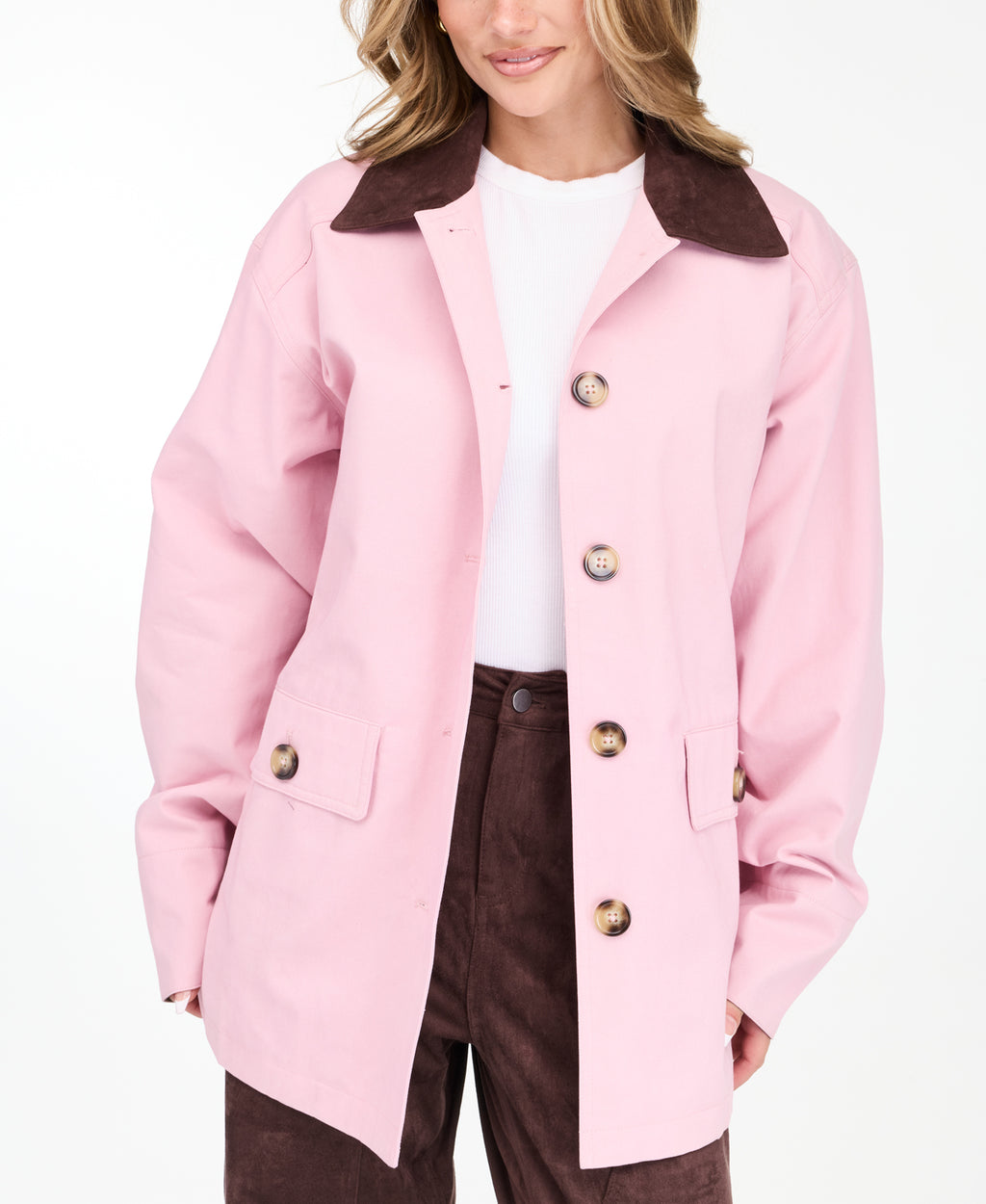 A woman modeling a light pink oversized jacket with dark brown collar and buttons, worn over a white top and paired with a brown pants, on a white studio background.