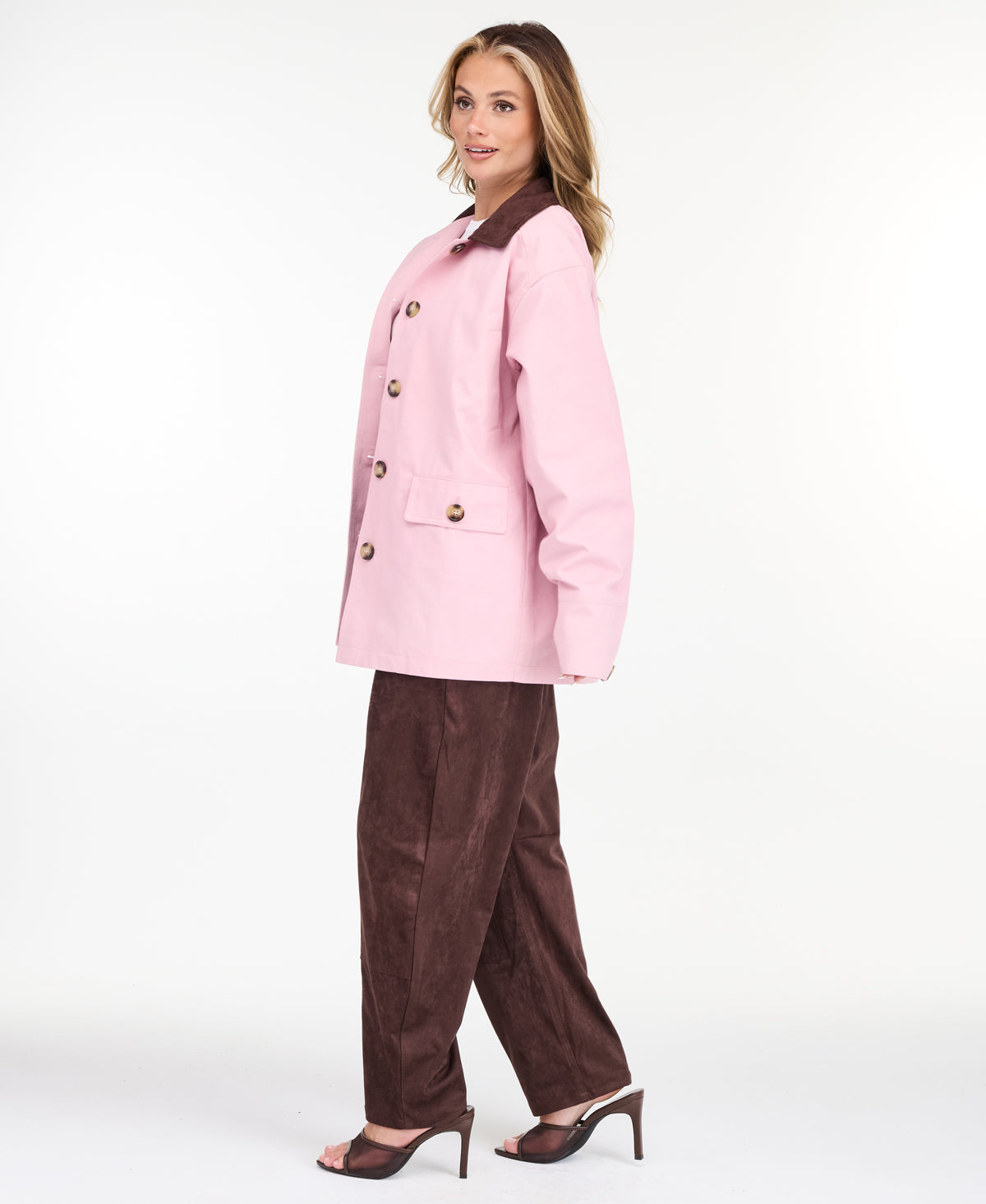 A woman modeling a light pink oversized jacket with dark brown collar and buttons, worn over a white top and paired with a brown pants, on a white studio background.