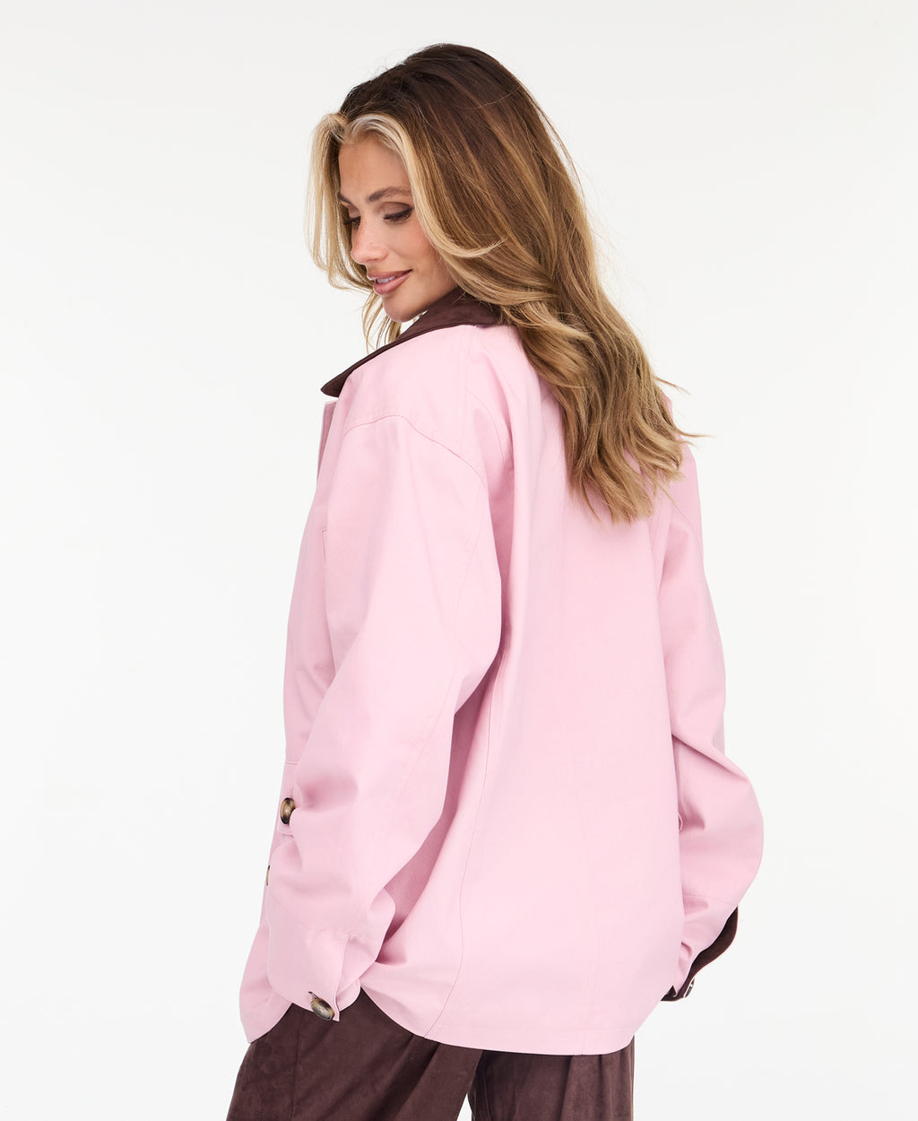 A woman modeling a light pink oversized jacket with dark brown collar and buttons, worn over a white top and paired with a brown pants, on a white studio background.
