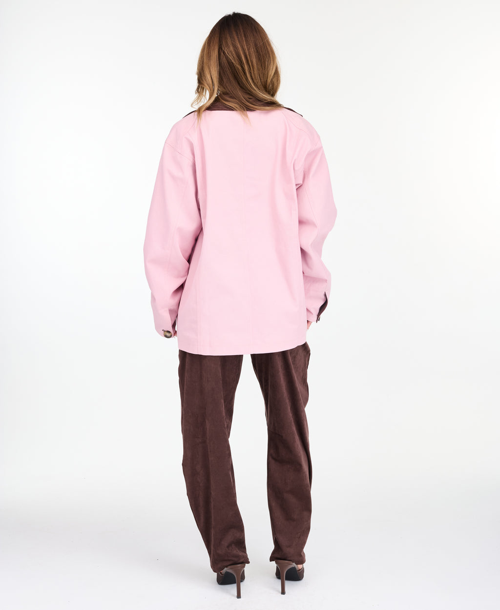 A woman modeling a light pink oversized jacket with dark brown collar and buttons, worn over a white top and paired with a brown pants, on a white studio background.