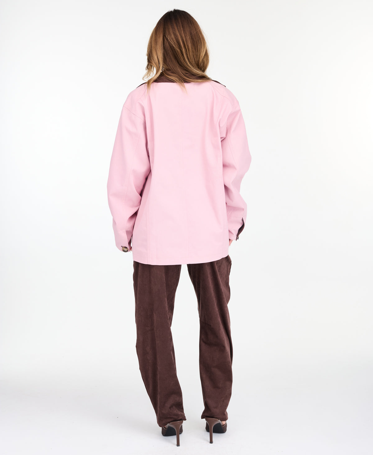 A woman modeling a light pink oversized jacket with dark brown collar and buttons, worn over a white top and paired with a brown pants, on a white studio background.