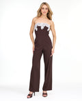 A woman modeling a dark brown pinstriped strapless jumpsuit with a white floral lace bodice trim, photographed against a plain white studio background.