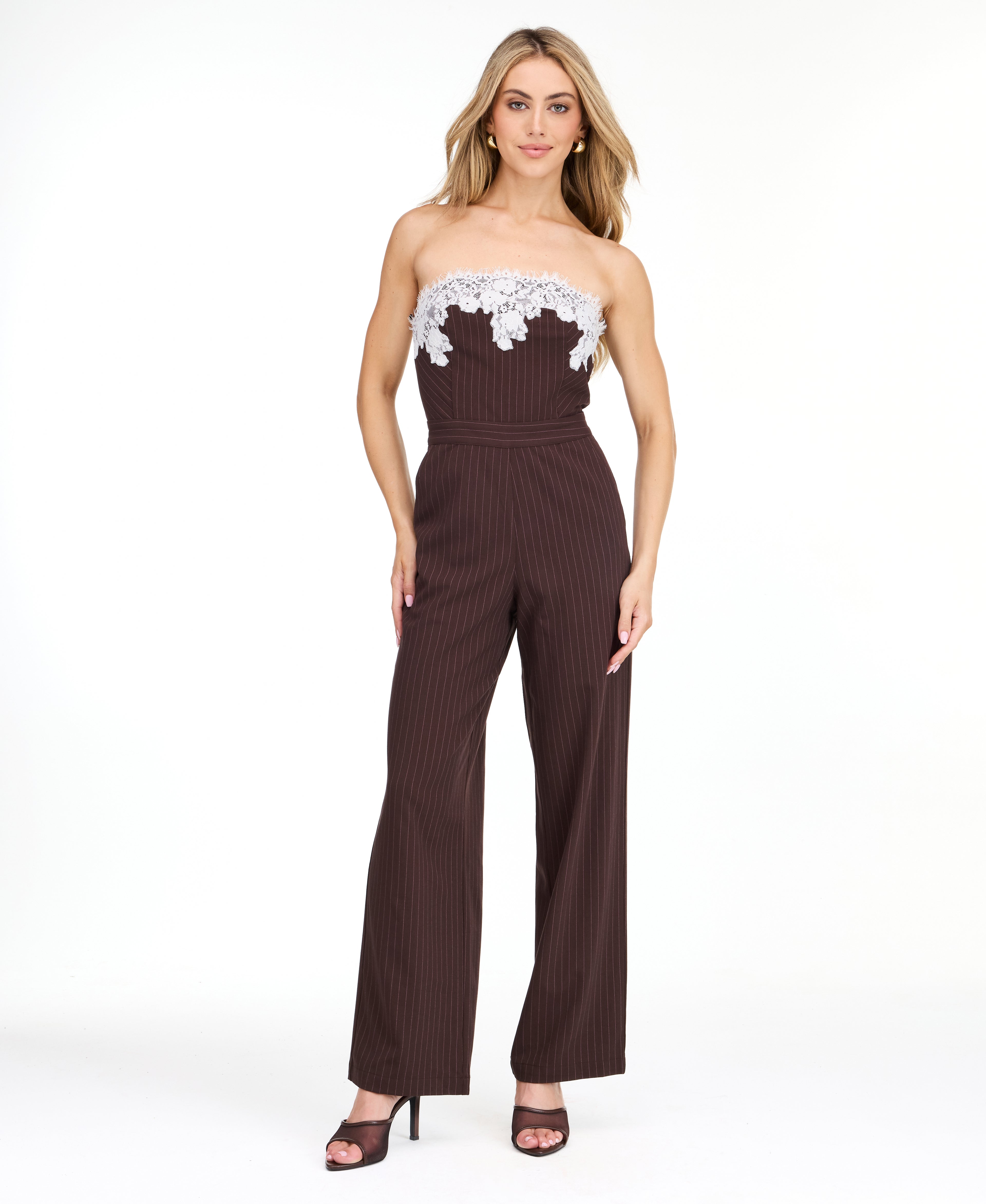 A woman modeling a dark brown pinstriped strapless jumpsuit with a white floral lace bodice trim, photographed against a plain white studio background.