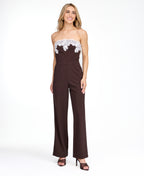 A woman modeling a dark brown pinstriped strapless jumpsuit with a white floral lace bodice trim, photographed against a plain white studio background.
