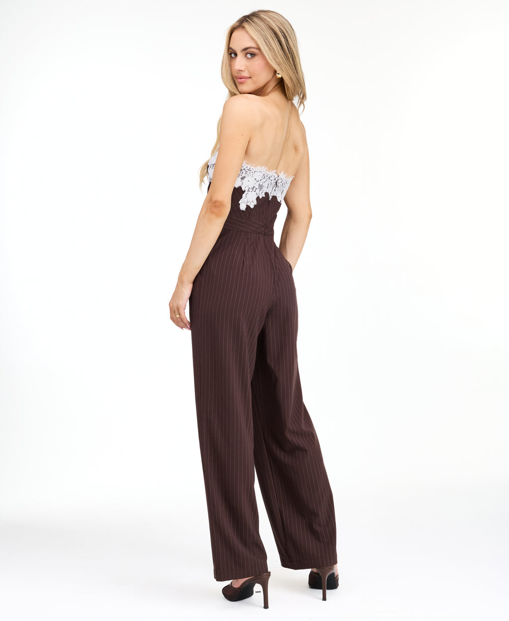 A woman modeling a dark brown pinstriped strapless jumpsuit with a white floral lace bodice trim, photographed against a plain white studio background.