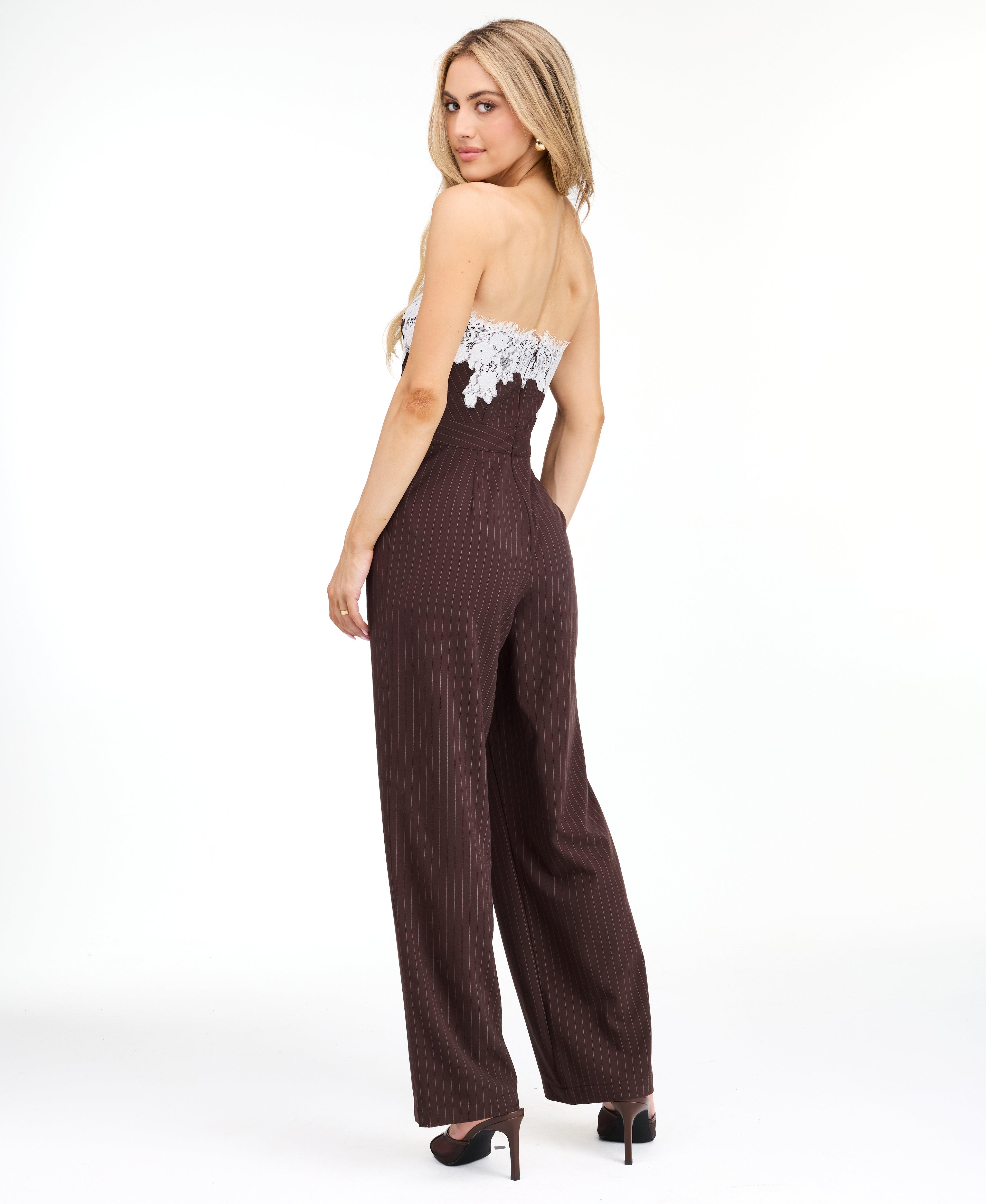 A woman modeling a dark brown pinstriped strapless jumpsuit with a white floral lace bodice trim, photographed against a plain white studio background.