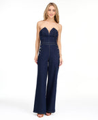 Model wearing a strapless  dark wash denim jumpsuit with heels standing against a white background
