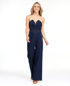 Model wearing a strapless  dark wash denim jumpsuit with heels standing against a white background