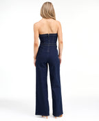 Model wearing a strapless  dark wash denim jumpsuit with heels standing against a white background
