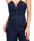 Model wearing a strapless  dark wash denim jumpsuit with heels standing against a white background