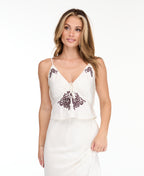 A woman modeling a white tank with matching midi skirt with dark brown floral lace on the bodice and hem, set against a plain white studio background.
