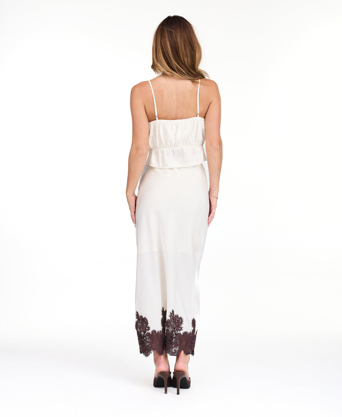 A woman modeling a white tank with matching midi skirt with dark brown floral lace on the bodice and hem, set against a plain white studio background.