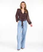 Model wearing a brown chiffon polka dot top with a removable neck scarf, medium wash denim jeans, and brown kitten heels
