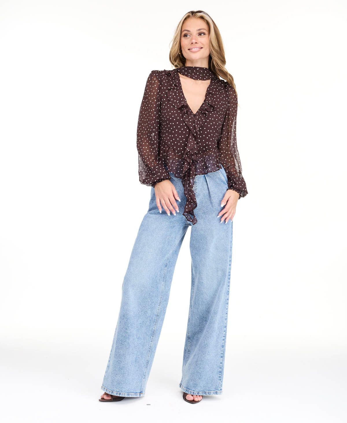 Model wearing a brown chiffon polka dot top with a removable neck scarf, medium wash denim jeans, and brown kitten heels
