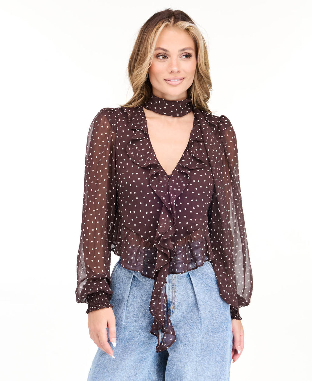 Model wearing a brown chiffon polka dot top with a removable neck scarf, medium wash denim jeans, and brown kitten heels