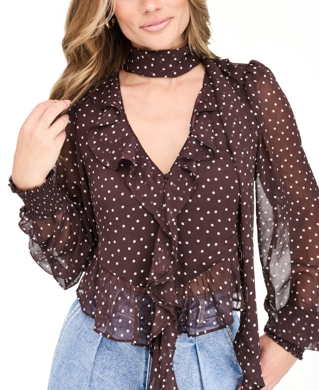 Model wearing a brown chiffon polka dot top with a removable neck scarf, medium wash denim jeans, and brown kitten heels