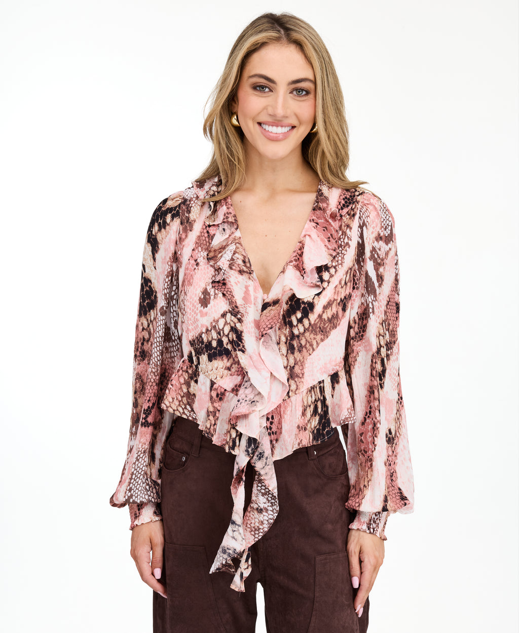 Model wearing a pink snake print top with brown suede pants and brown kitten heels standing against a white back drop