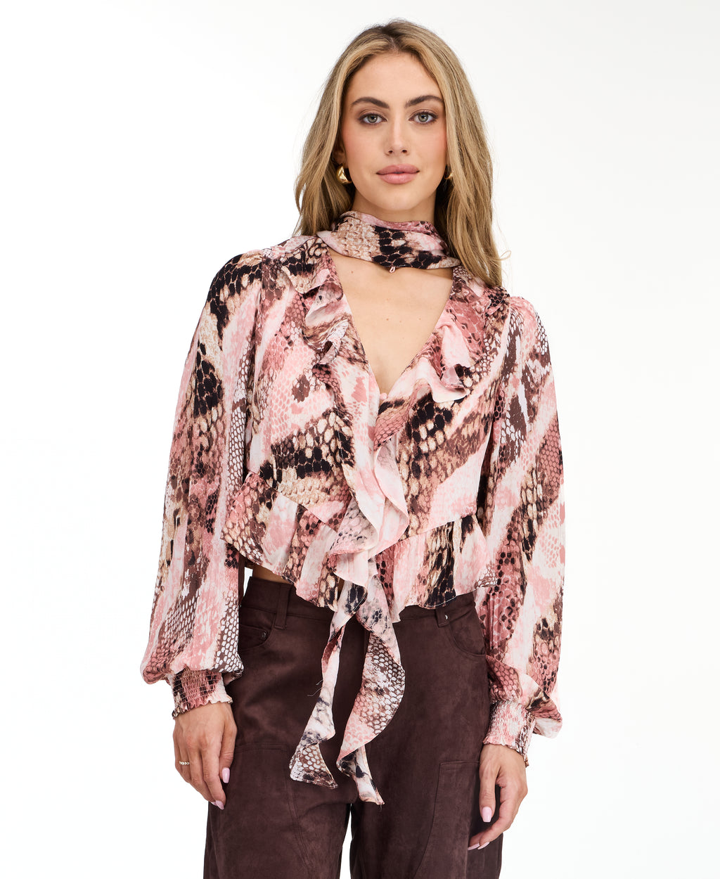 Model wearing a pink snake print top with brown suede pants and brown kitten heels standing against a white back drop