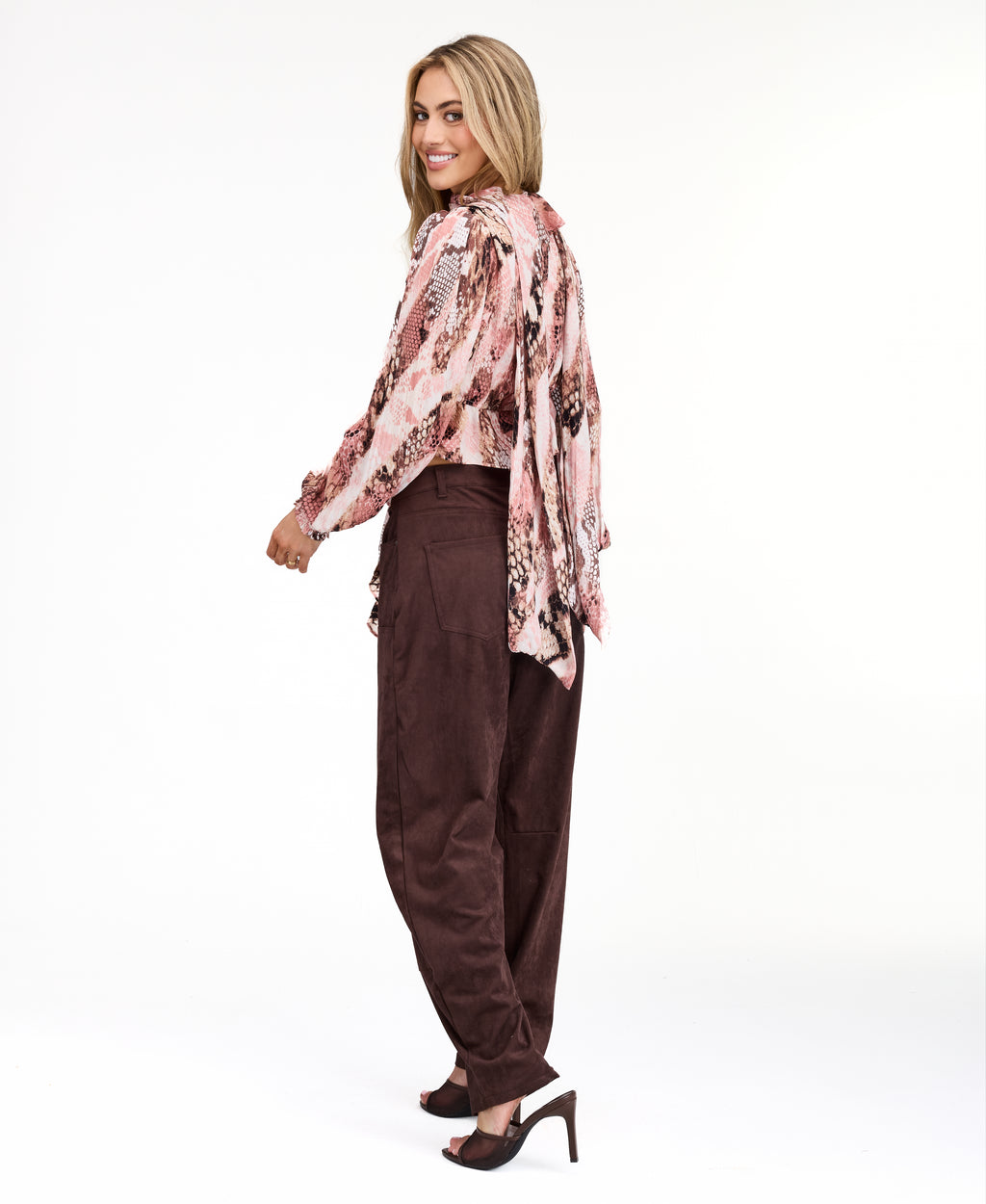 Model wearing a pink snake print top with brown suede pants and brown kitten heels standing against a white back drop