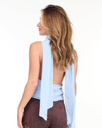 Model wearing a blue cowl neck tank with a removable neck scarf paired with brown sueded pants standing on a white background