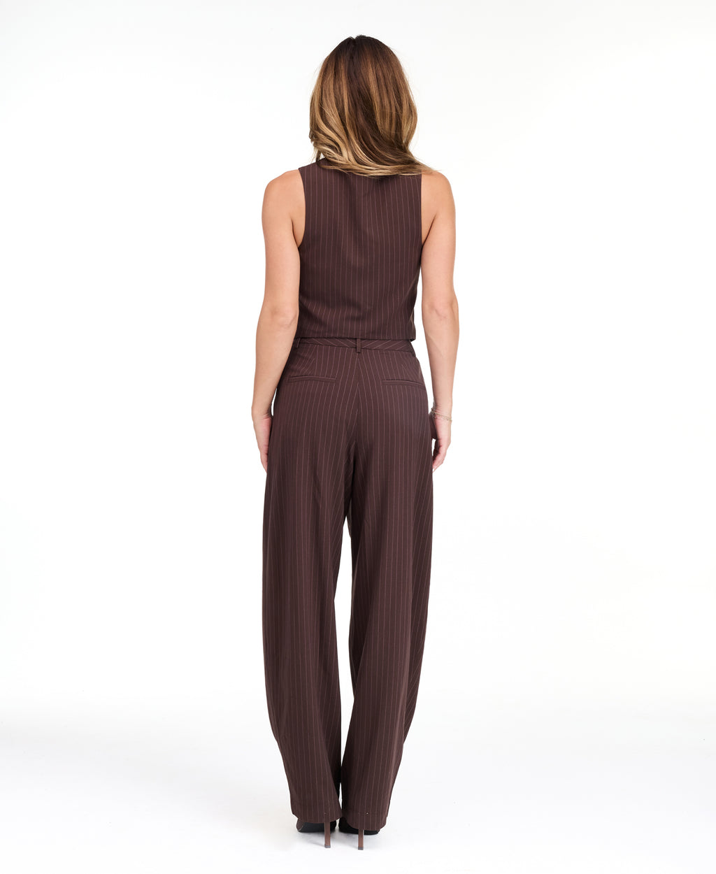 A woman modeling a dark brown pinstriped sleeveless vest-style top and barrel pants, standing against a plain white studio background.