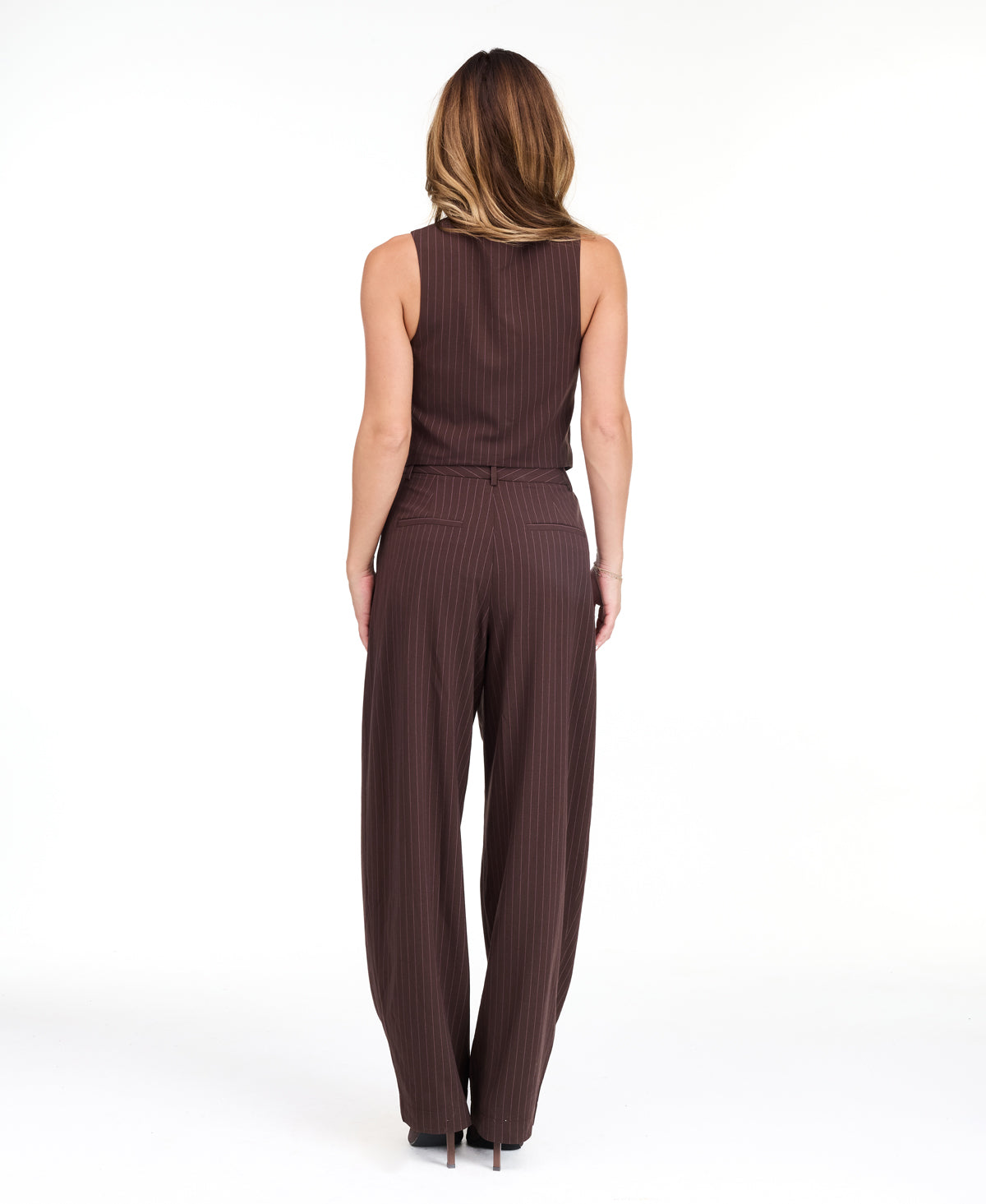 A woman modeling a dark brown pinstriped sleeveless vest-style top and barrel pants, standing against a plain white studio background.