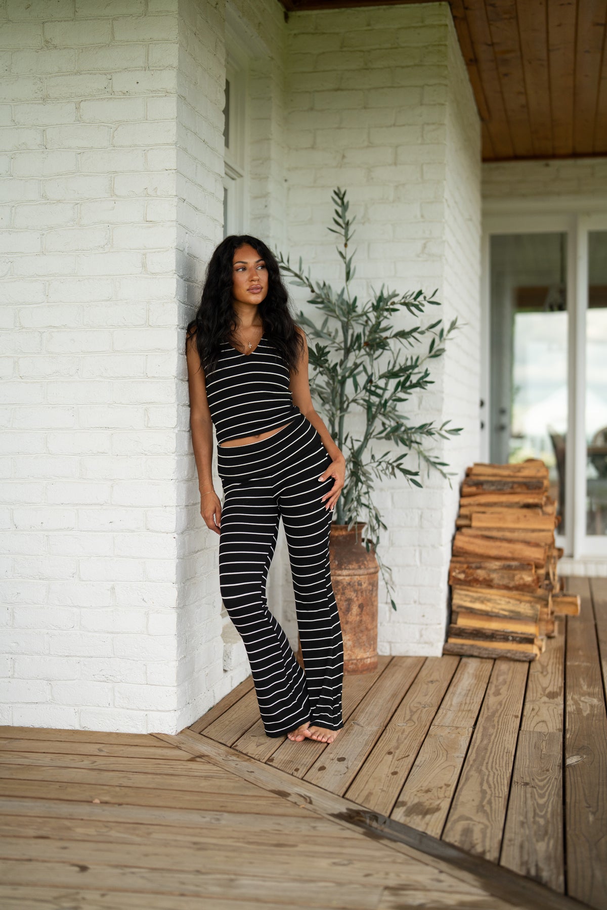 A woman stands barefoot on a wooden porch wearing a matching black and white horizontally striped crop top and flared pants set. The setting features a white brick wall, a potted olive tree, and a stack of firewood in the background.