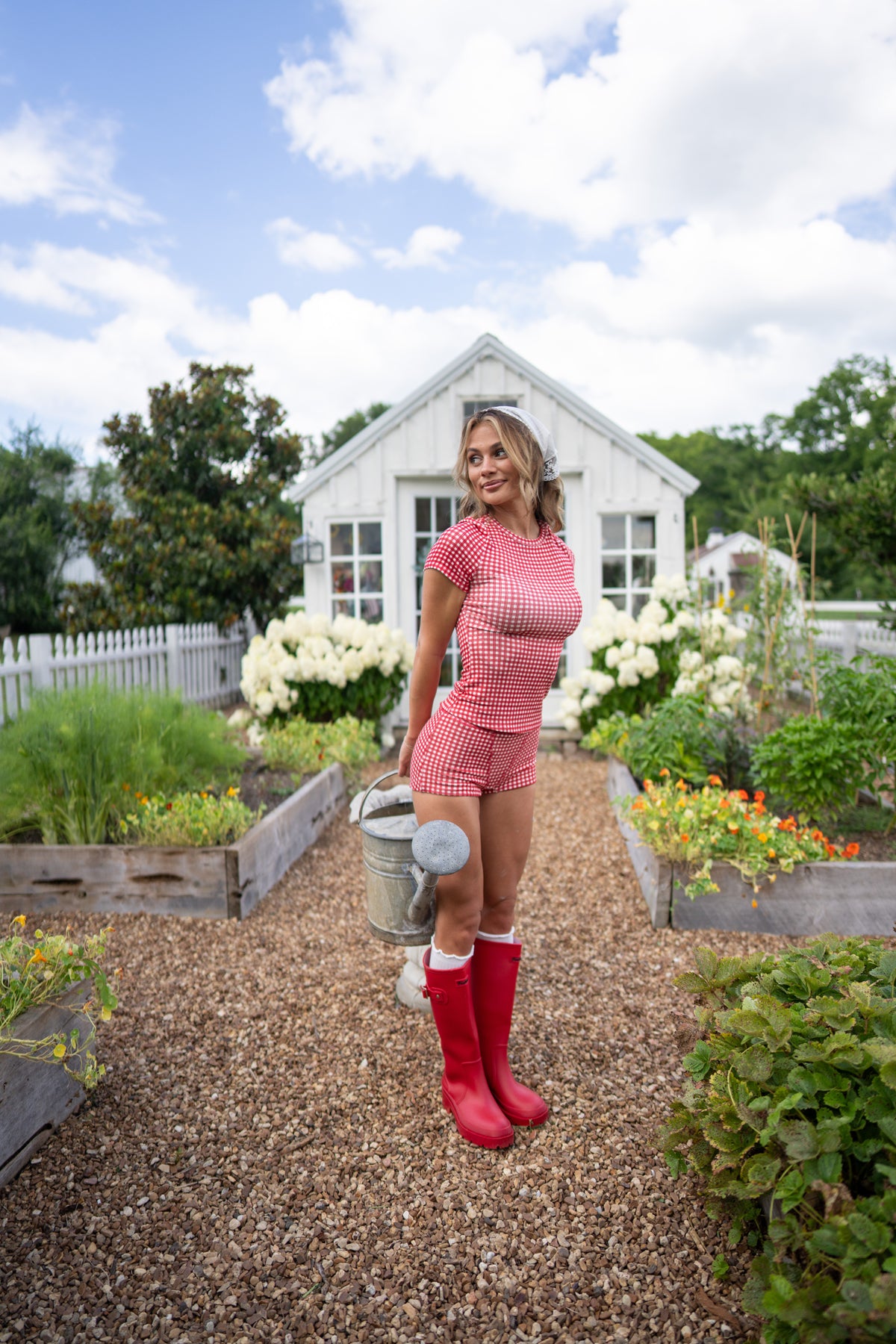 A woman stands in a garden wearing a red-and-white gingham short-sleeve top and matching shorts, red rain boots, and holding a metal watering can. A white garden shed, raised garden beds, and a blue sky are in the background.