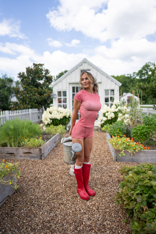 A woman stands in a garden wearing a red-and-white gingham short-sleeve top and matching shorts, red rain boots, and holding a metal watering can. A white garden shed, raised garden beds, and a blue sky are in the background.