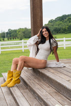 A woman sits on rustic wooden steps outdoors wearing a white patterned bodysuit, a white cardigan, and bright yellow rain boots, with a grassy field and white fence in the background.