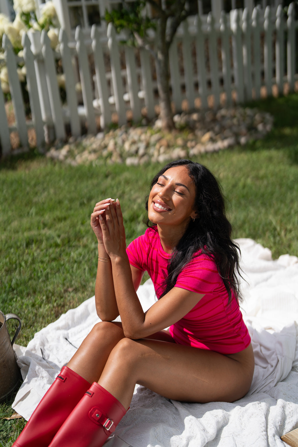 A woman sitting outdoors on a white blanket in a backyard, wearing a bright pink short-sleeve top and red rain boots.