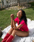 A woman sitting outdoors on a white blanket in a backyard, wearing a bright pink short-sleeve top and red rain boots.