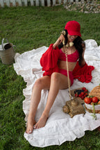 A woman sitting outdoors on a white picnic blanket on grass, wearing a bright red bikini and red cap while holding a vintage-style telephone. A basket of fruit and a metal bucket are nearby.