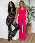 Two women modeling matching ribbed lounge sets in a bright indoor setting with a white brick wall and wooden floor. One set is black and white striped, and the other is hot pink stripes.