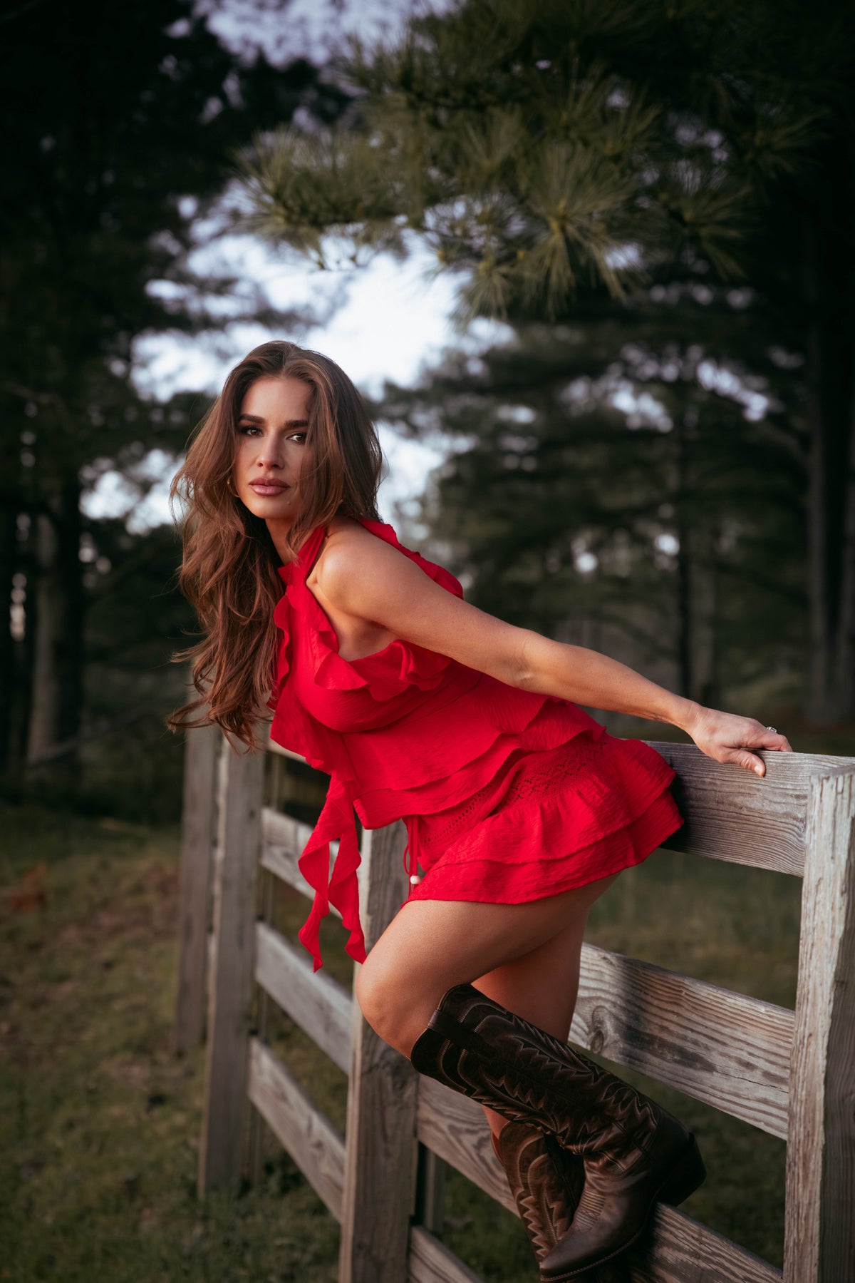 Jessie in a red skirt and top and brown boots leaning against a wooden fence